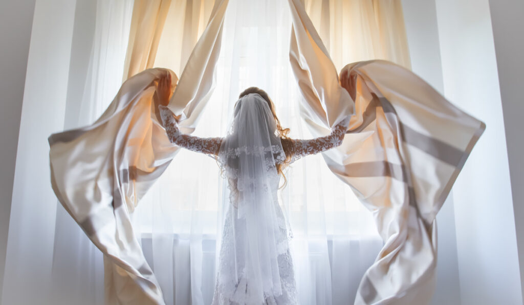Back view long hair bride in white wedding dress veil open window curtains