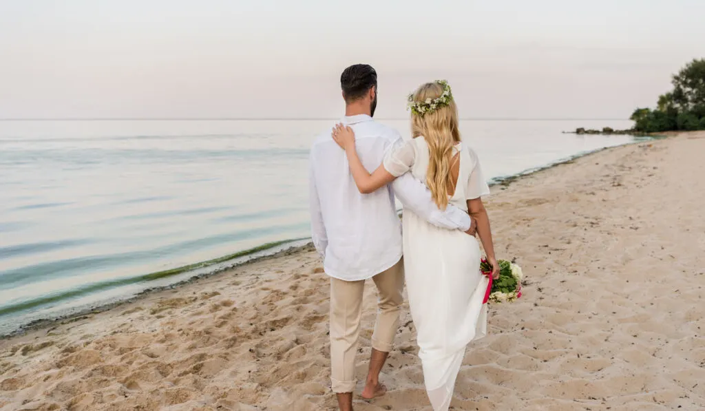 rear view of bride with wedding bouquet and groom hugging and walking on beach