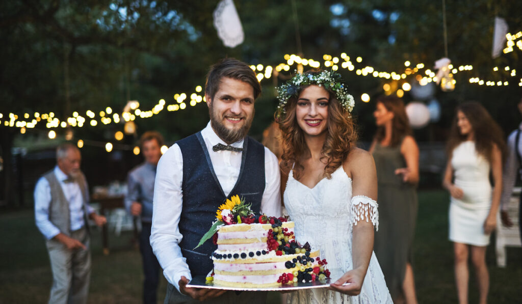 Bride and groom holding a cake at wedding reception outside in the backyard.
