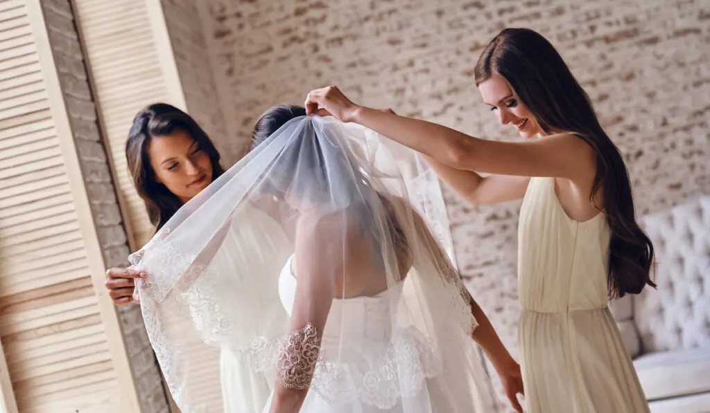  Bridesmaids helping bride to put on a veil in the fitting room