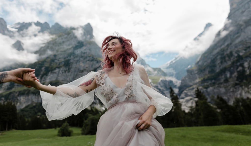 Happy Bride in Wedding Dress Holding Hands With Groom on Meadow With Mountains and Clouds in Alps 