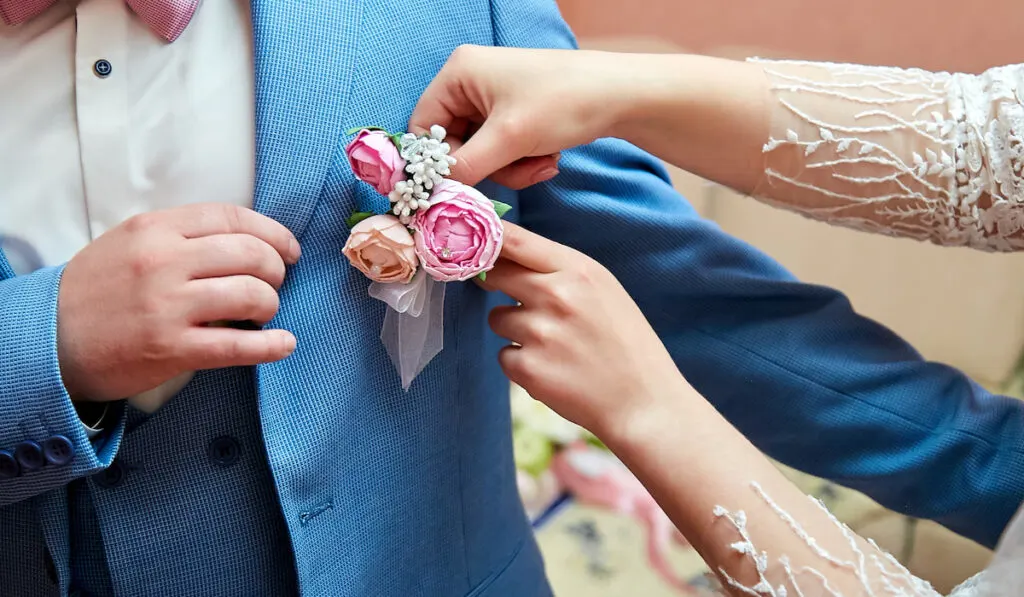 The bride's hand puts on a boutonniere flower on the groom's jacket.
