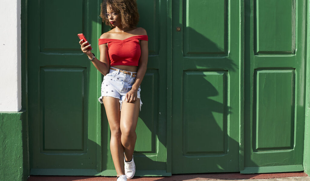 Woman in red top and shorts standing on green door checking her phone