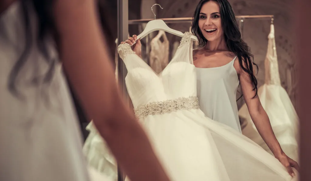 Woman  is looking into the mirror and smiling while choosing elegant wedding dresses in modern wedding salon