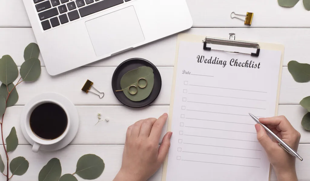 woman holding a pen working on her wedding checklist. With her laptop, cup of coffee and wedding rings on the table.