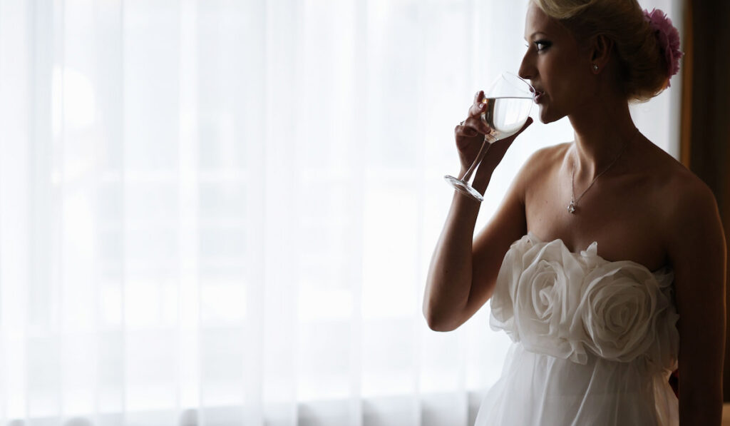bride drinking water in front of a window 