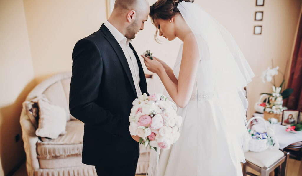 bride on her wedding dress with short veil putting stylish boutonniere on groom suit 