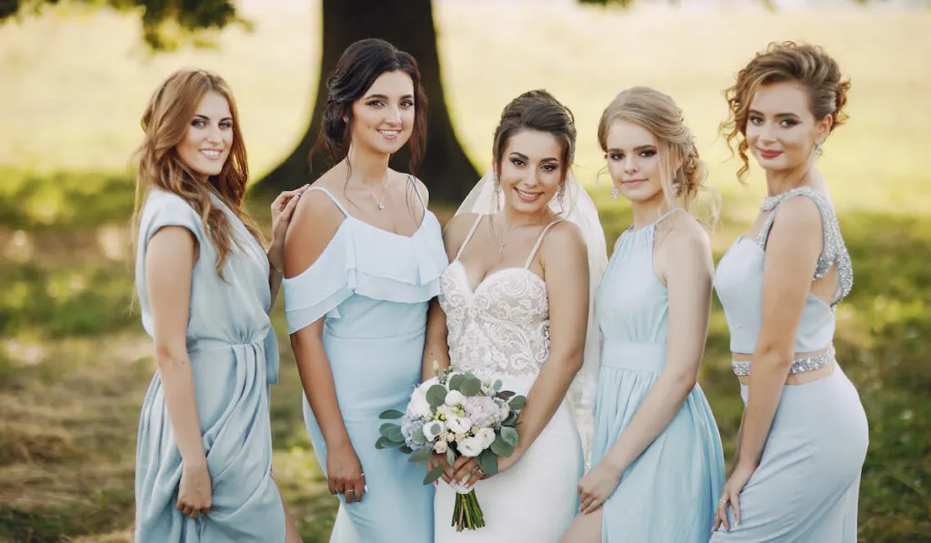 bride wearing white wedding dress along with her bridesmaids in blue dresses standing in a park