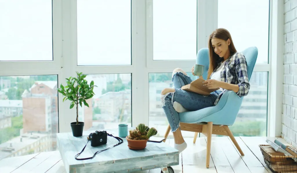 woman relaxing at home, reading book and drinking coffee sitting on balcony in apartment 