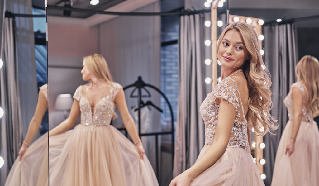 woman wearing pink wedding dress and smiling while standing in front of the mirror in bridal shop