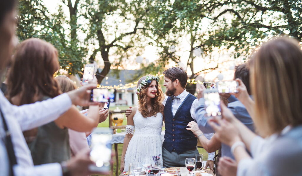Guests with smartphones taking photo of bride and groom at wedding reception outside