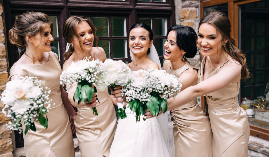 A bride in a wedding dress and bridesmaids in golden dresses hold stylish bouquets of flowers on their wedding day