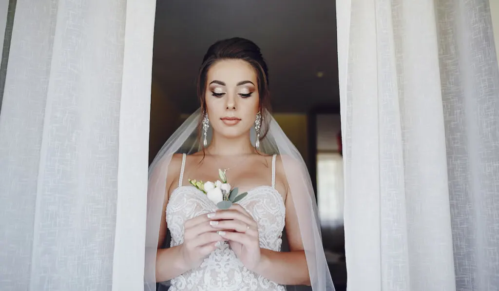 A young and beautiful bride at home standing near window with flowers

