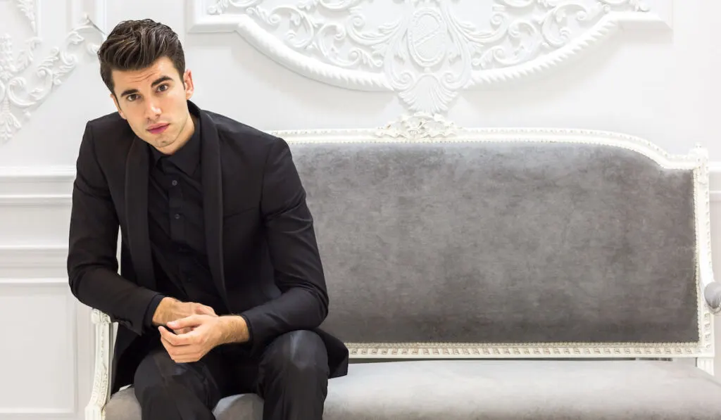 Elegant young groom  wearing all black suit sitting in the sofa posing at the camera