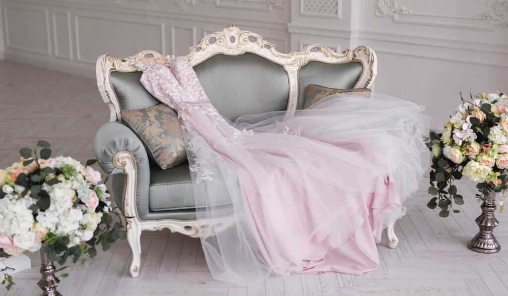 Luxurious pink wedding dress is lying on a grey sofa in a white room

