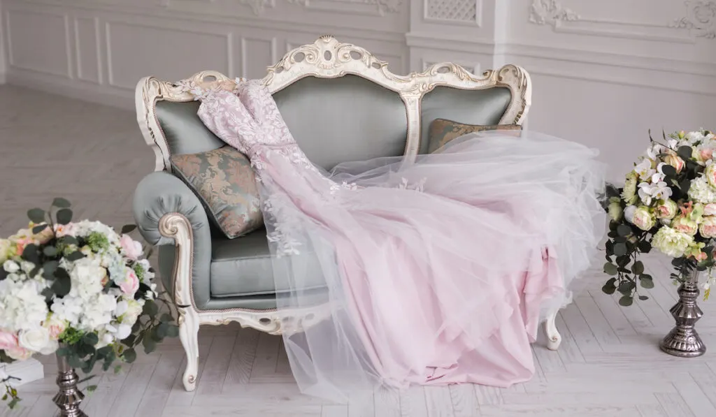 Luxurious pink wedding dress is lying on a grey sofa in a white room
