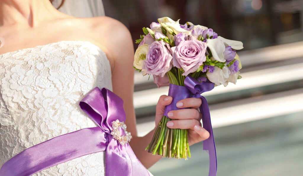 bride wearing white wedding dress with purple ribbon and holding purple bouquet of flowers