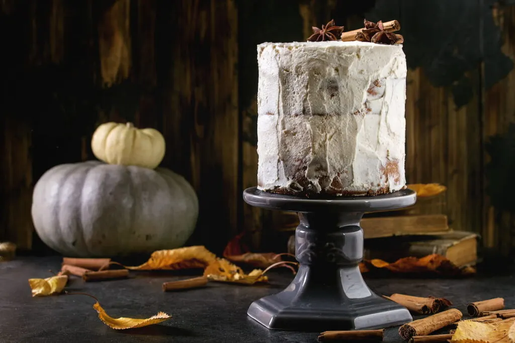 white naked cake decorated by cinnamon sticks and anise on cake stand with yellow leaves and decorative pumpkins above on black table, Dark rustic style cake 