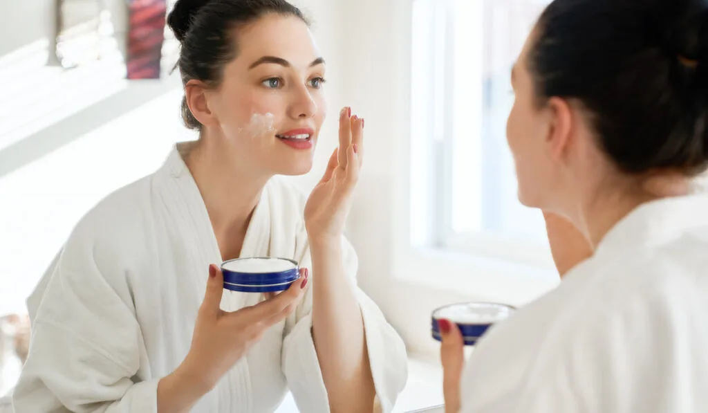 woman applying cream on her face looking in the mirror in the bathroom