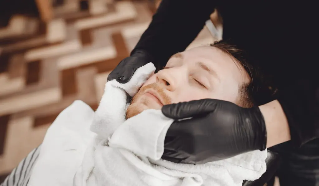 Man sits in barbershop chair and steam his face with hot towel