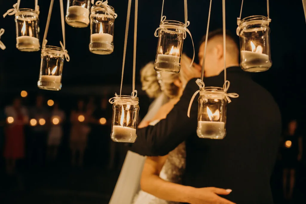 small candles in a clear jars hanging outside the wedding venue, bride and groom in the background